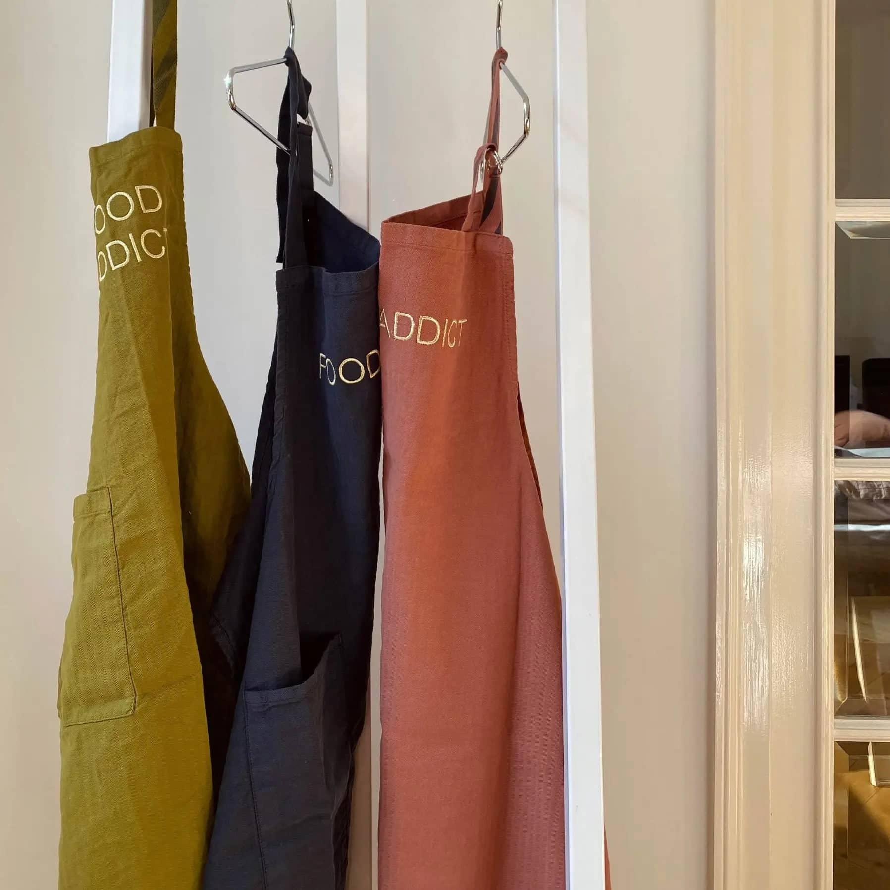 Collection de tablier en lin suspendus en vert, noir et terracotta avec poche et inscription food addict